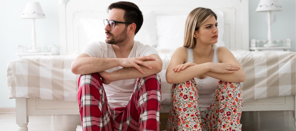 a man and a woman in pajamas sit next to each other, looking away from one another and clearly upset
